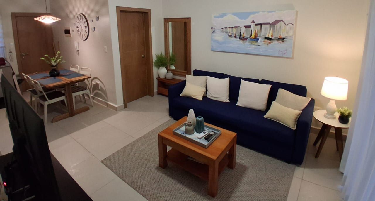Een woonkamer met een blauwe bank, een houten salontafel, een eettafel met stoelen, een lamp en een schilderij aan de muur.