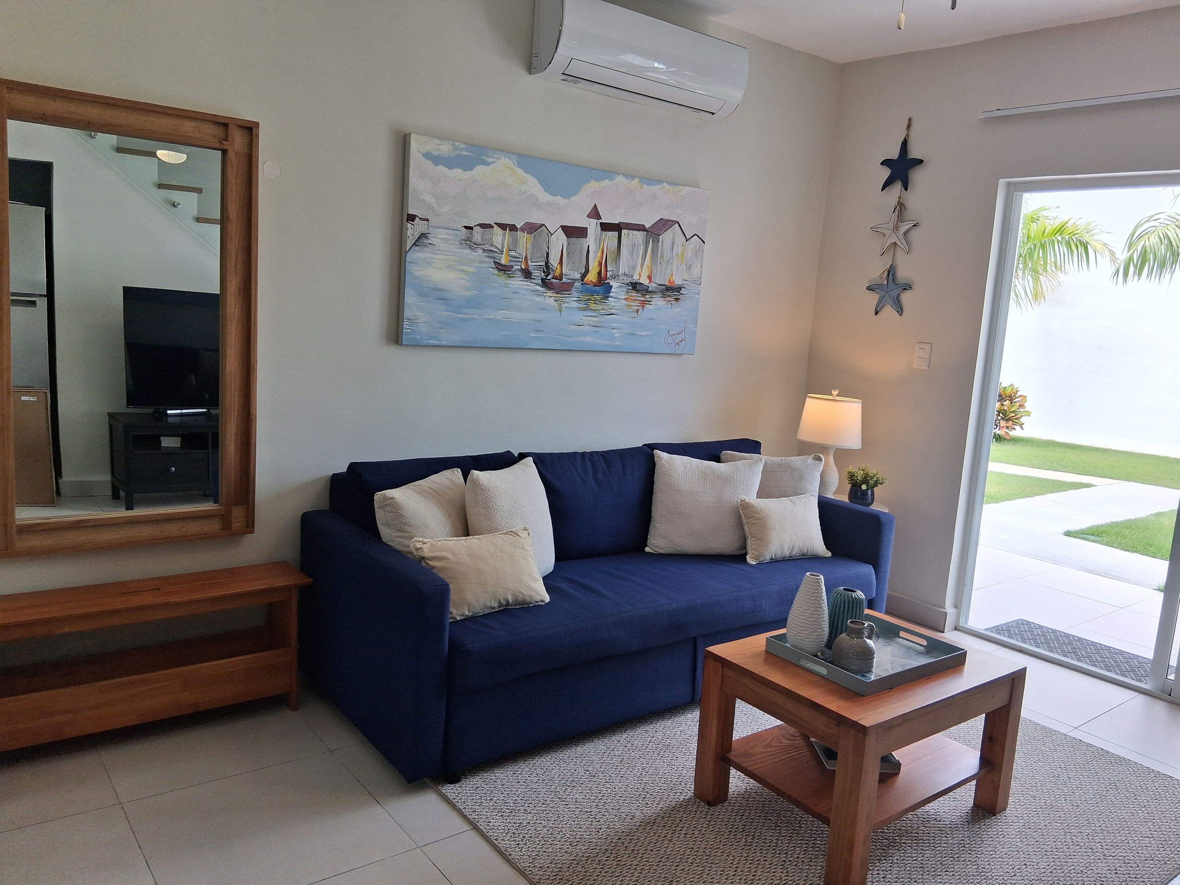A living room with a blue sofa, a coffee table, a painting of boats, a star decoration, and a large window.