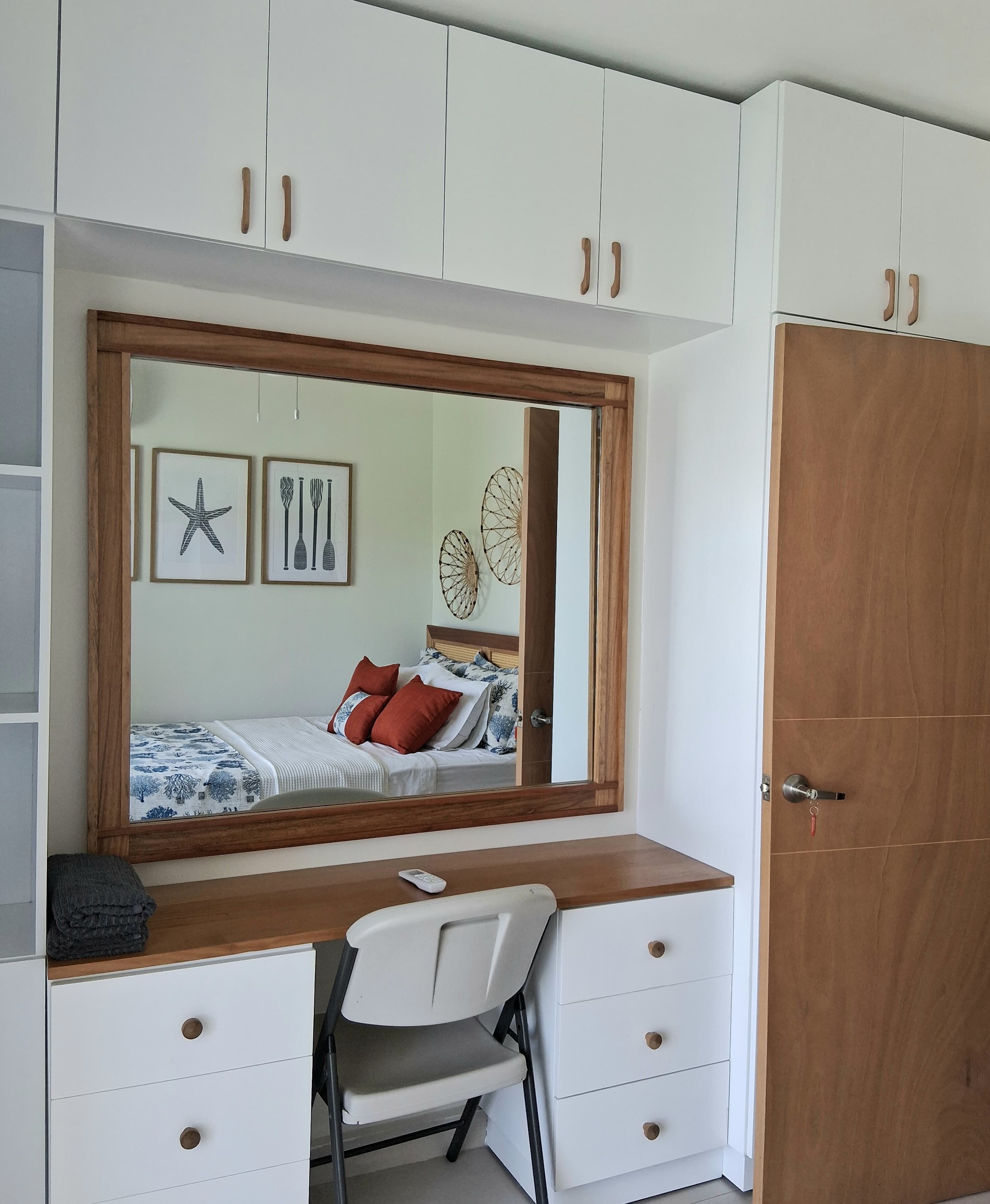 A bedroom with a large mirror, a bed with a white and blue patterned bedspread, a wooden desk with a chair, and a wardrobe with white doors.
