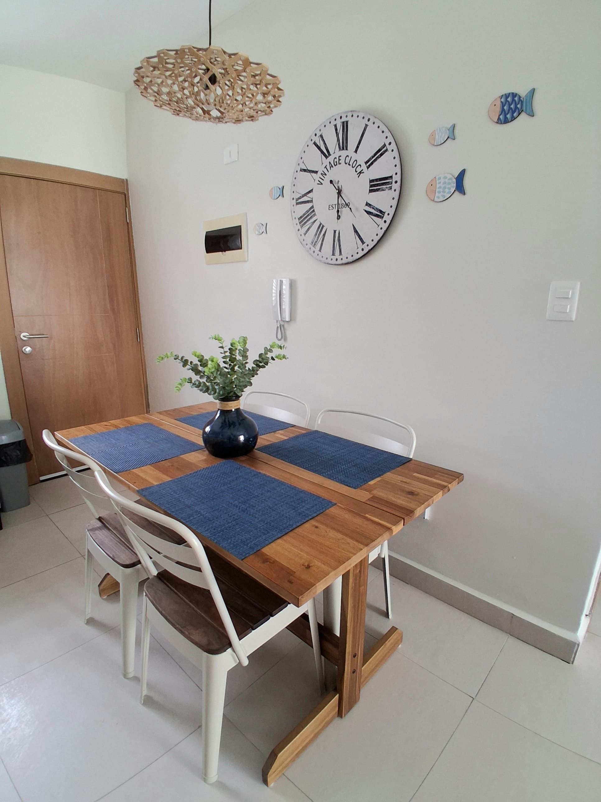 A dining area with a wooden table, white chairs, blue placemats, a decorative vase, and a wall-mounted vintage clock.