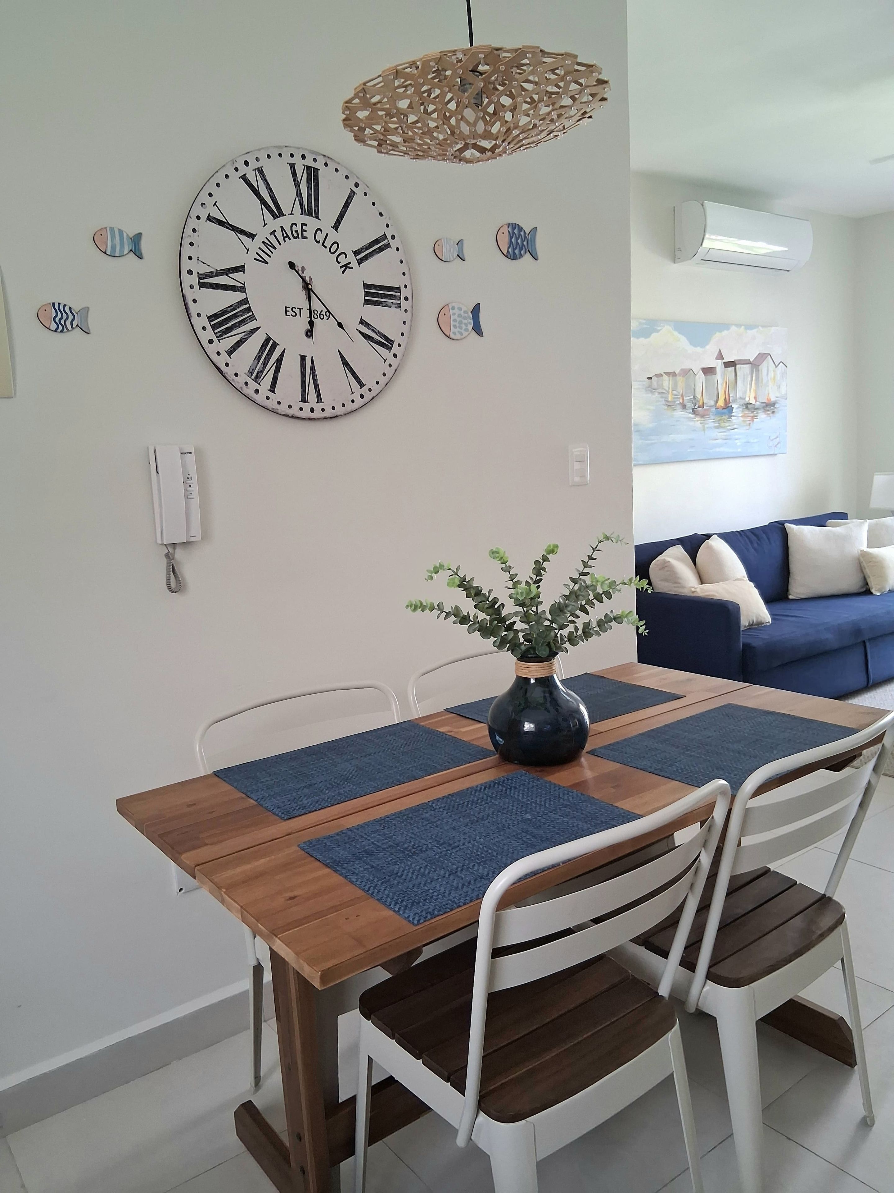 Un comedor con mesa de madera, sillas blancas, un jarrón azul con follaje y un cuadro de peces decorativo.