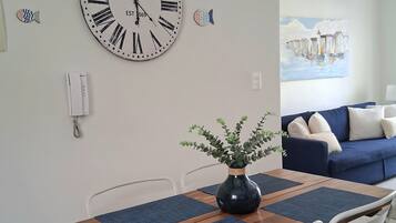 Un comedor con mesa de madera, sillas blancas, un jarrón azul con follaje y un cuadro de peces decorativo.