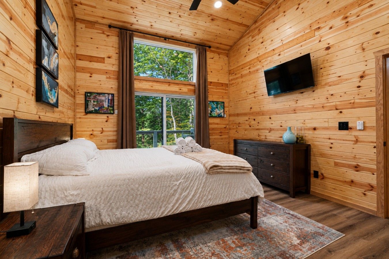 A wooden-paneled bedroom with a bed, a dresser, a TV, and artwork on the walls.