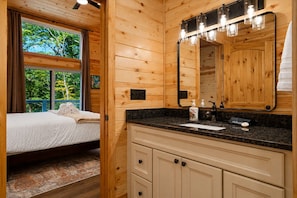 Una habitación de cabaña de madera con una cama, un baño con lavamanos y espejo, y una ventana con vistas a árboles.