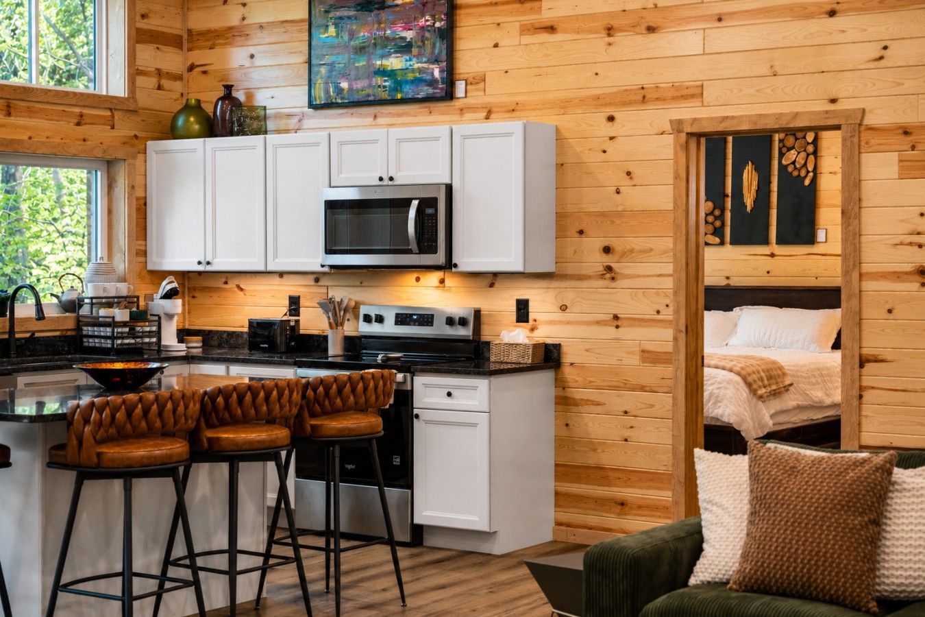 Cocina compacta con armarios blancos, microondas y estufa. Hay dos taburetes de bar y una pequeña mesa de comedor. Se ve una cama con ropa blanca de cama al fondo.