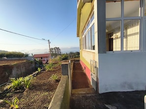 A building with a small garden area in front, a clear sky, and a distant view of a red-roofed house.
