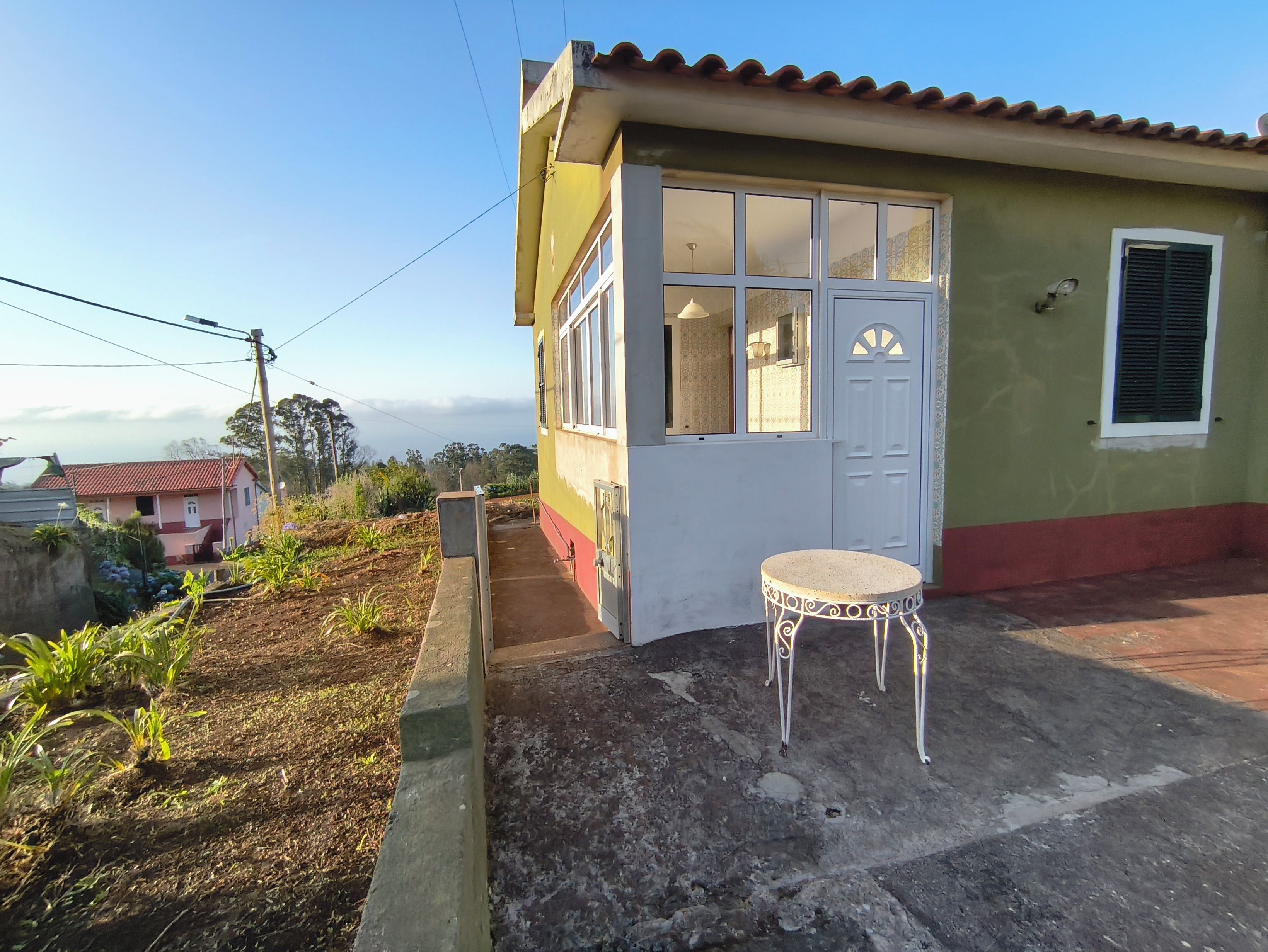 A small building with a door and windows, a small table, and a garden area with plants.