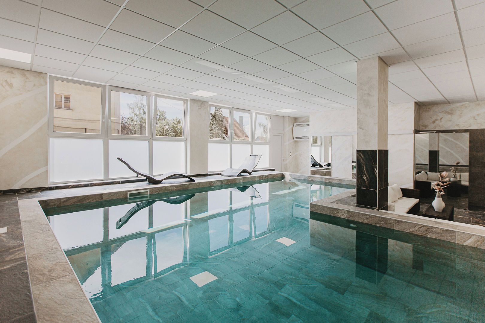 Una piscina interior con agua cristalina, rodeada por un suelo y paredes de baldosas, con ventanas amplias que permiten la entrada de luz natural.