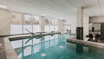 Una piscina interior con agua cristalina, rodeada por un suelo y paredes de baldosas, con ventanas amplias que permiten la entrada de luz natural.