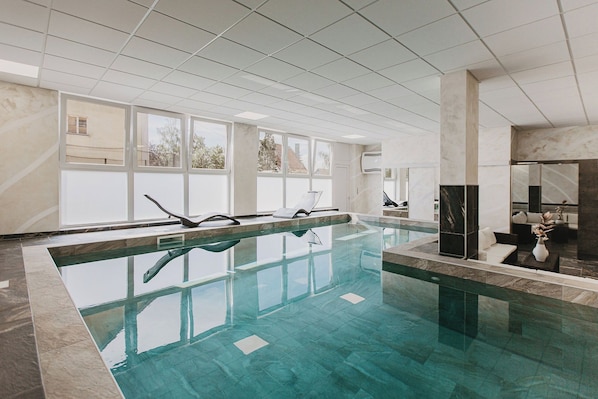 Una piscina interior con agua cristalina, rodeada por un suelo y paredes de baldosas, con ventanas amplias que permiten la entrada de luz natural.