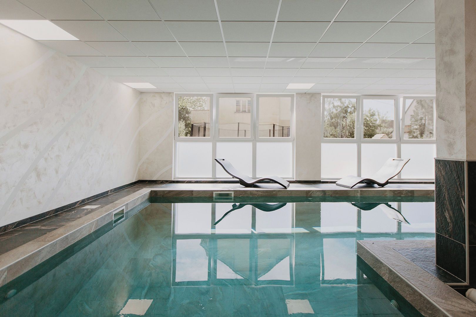 Una piscina interior con dos sillas reclinables, ventanas amplias y techo de azulejos.