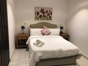 A hotel room with a bed, two bedside tables, a headboard, a wall-mounted painting, and a folded towel on the bed. - The Lennox Apartments-Airport (Accra)