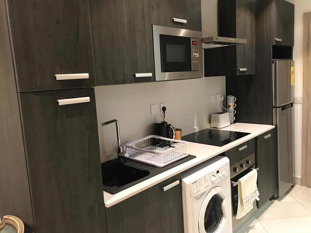 A modern kitchen with dark cabinetry, a built-in microwave, a stainless steel refrigerator, a washing machine, and a countertop with a dish rack.