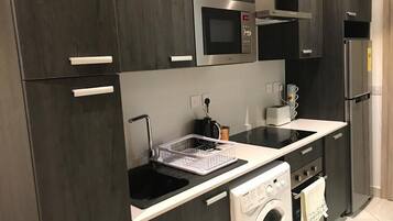 A modern kitchen with dark cabinetry, a built-in microwave, a stainless steel refrigerator, a washing machine, and a countertop with a dish rack.