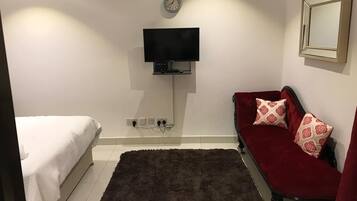 A bedroom with a bed, a television mounted on the wall, a red sofa with patterned cushions, and a dark rug on the floor.