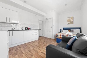 A modern kitchen with white cabinets and a wooden floor, a bed with a blue headboard, and a gray sofa. - Beautiful studio near Leeds City Centre - Room 6 (Leeds)