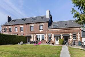 Un edificio de ladrillo de dos plantas con techo plano, múltiples ventanas y un pequeño jardín con mobiliario exterior.