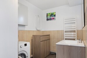 A compact bathroom with a washing machine, wooden cabinets, a sink with a mirror, and a towel rack. - Cottage 'L'ancienne Forge' with Private Terrace, Private Garden and Wi-Fi (Louvetot)