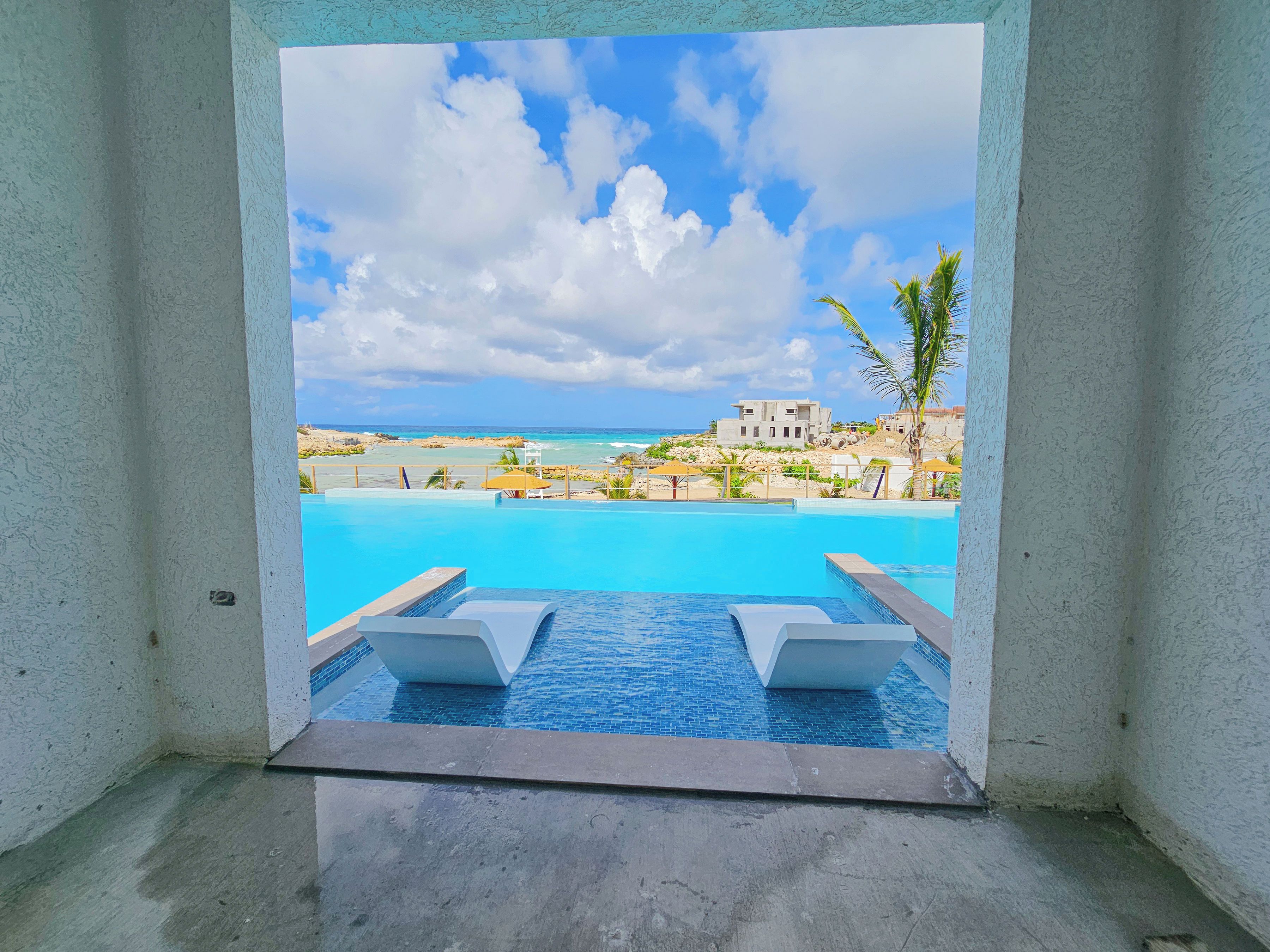 A clear view of a swimming pool with lounge chairs, a view of the beach, and a building in the distance.