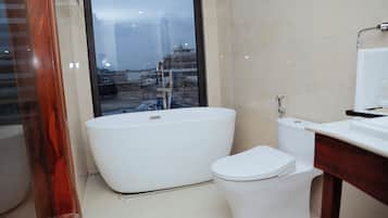 Een moderne badkamer met een vrijstaand bad, een toilet en een groot raam met uitzicht op een stadsbeeld.