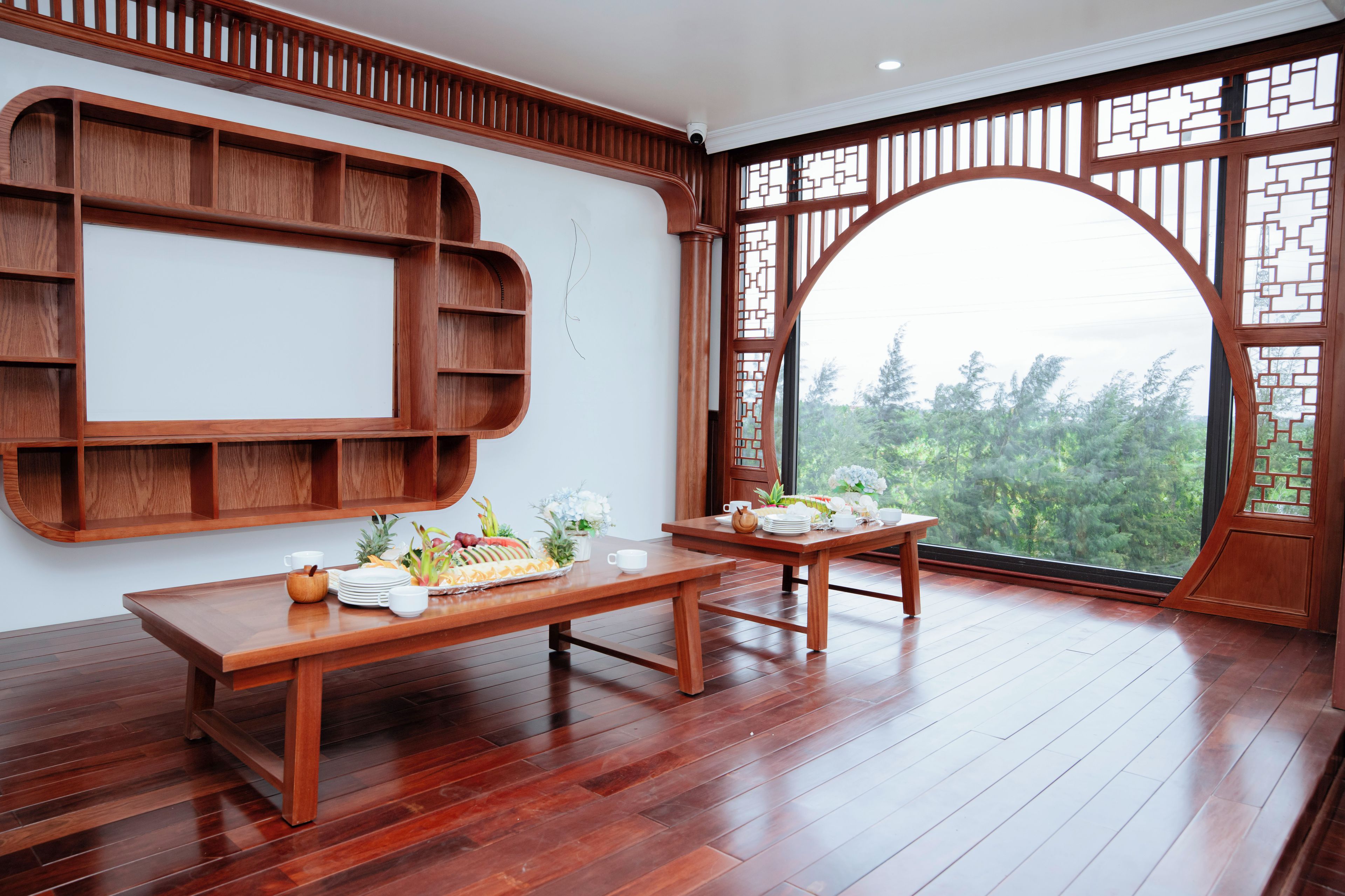 A room with a wooden dining table set with plates, cups, and a fruit platter. There is a wooden shelf unit against the wall and a large window with a view of trees.