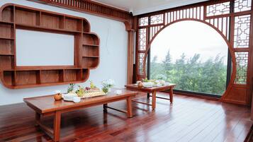 A room with a wooden dining table set with plates, cups, and a fruit platter. There is a wooden shelf unit against the wall and a large window with a view of trees.