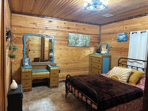 A wooden-paneled bedroom with a bed, dresser, vanity, and artwork.