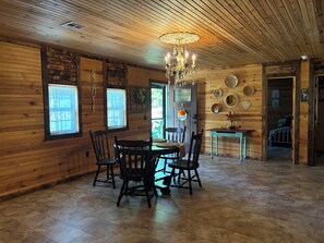 A wooden interior with a dining table, chairs, a chandelier, and a door leading to another room.