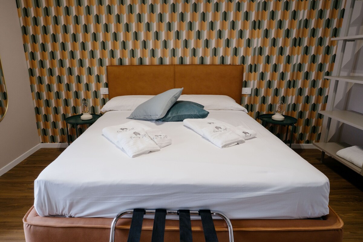 Een net opgemaakt bed met witte lakens, twee kussens en twee handdoeken met tekst, voor een behang met een patroon.
