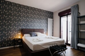 Een hotelkamer met een behang met patroon, een bed met witte lakens, een bedlamp en uitzicht op het omliggende landschap door een raam.