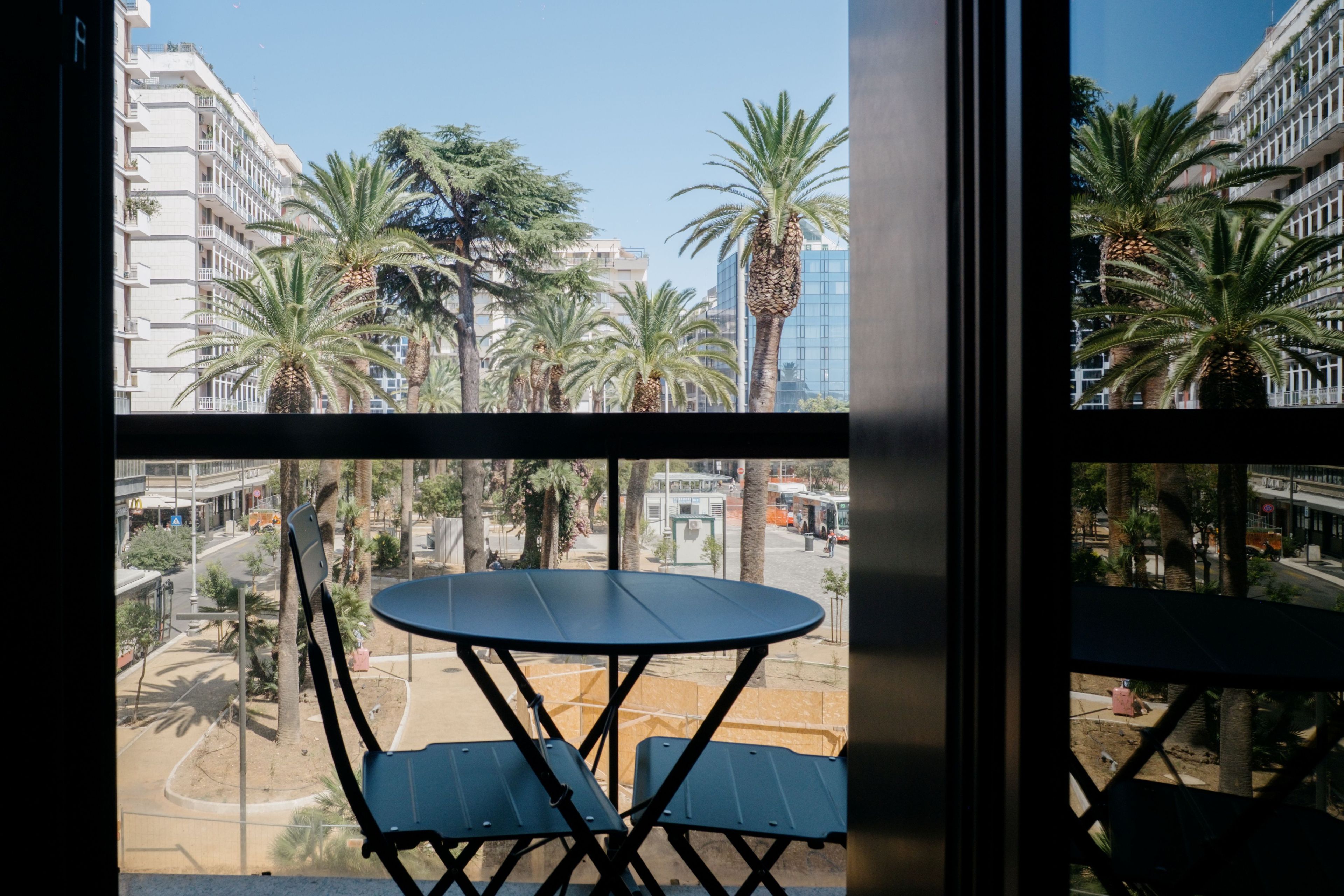 A balcony with a table and chairs, overlooking a street with palm trees and buildings.