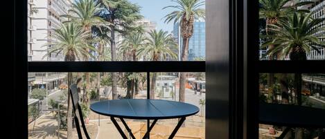 Un balcón con mesa y sillas, con vista a una calle con palmeras y edificios.