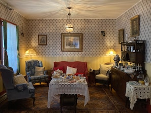 A room with patterned wallpaper, a red sofa, two armchairs, a table with a lace tablecloth, and a chandelier. - Room 3 - St. George Hotel (Barkerville)