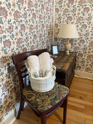 A room with floral wallpaper, a wooden chair with a cushion, a lamp, and a small table with a picture frame. - Room 3 - St. George Hotel (Barkerville)