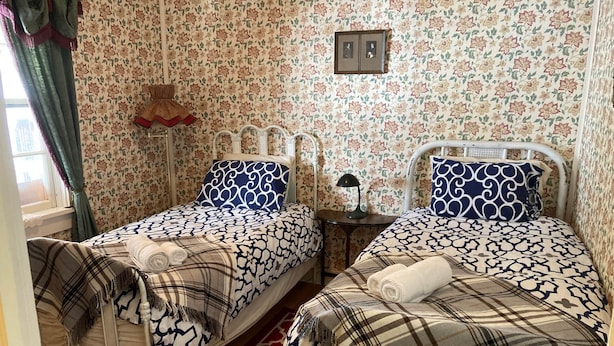 Quarto com duas camas, papel de parede floral, tapete vermelho estampado e janela com cortinas.