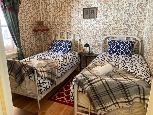 A bedroom with two beds, floral wallpaper, a red patterned rug, and a window with curtains. - Room 2 - St. George Hotel (Barkerville)