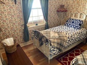 A bedroom with a bed, a window with curtains, a bedside table with a lamp, and a patterned wall. - Room 2 - St. George Hotel (Barkerville)