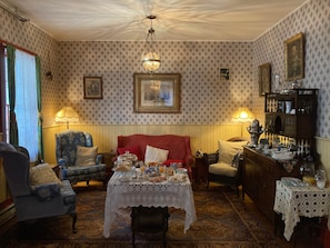 A room with patterned wallpaper, a red sofa, two armchairs, a table with a lace tablecloth, and a chandelier. - Room 2 - St. George Hotel (Barkerville)