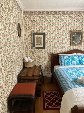 A bedroom with floral wallpaper, a wooden bed, a small bench, a wicker basket, and a framed picture on the wall.