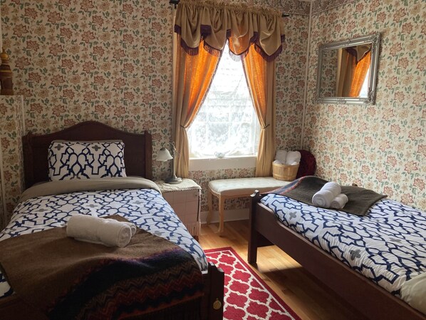 A bedroom with two beds, a bedside table, a lamp, a window with curtains, and a mirror on the wall. - Room 5 - St. George Hotel (Barkerville)