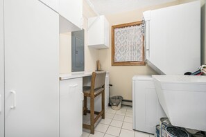 A compact laundry room with a washing machine, a drying rack, a chair, and a small trash bin. - On A Whim (Swanton)