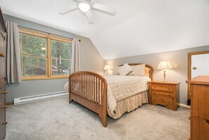 Une chambre avec un plafond en pente, un lit doté d’une tête de lit en bois, deux tables de chevet avec des lampes, une commode et une fenêtre avec des rideaux.