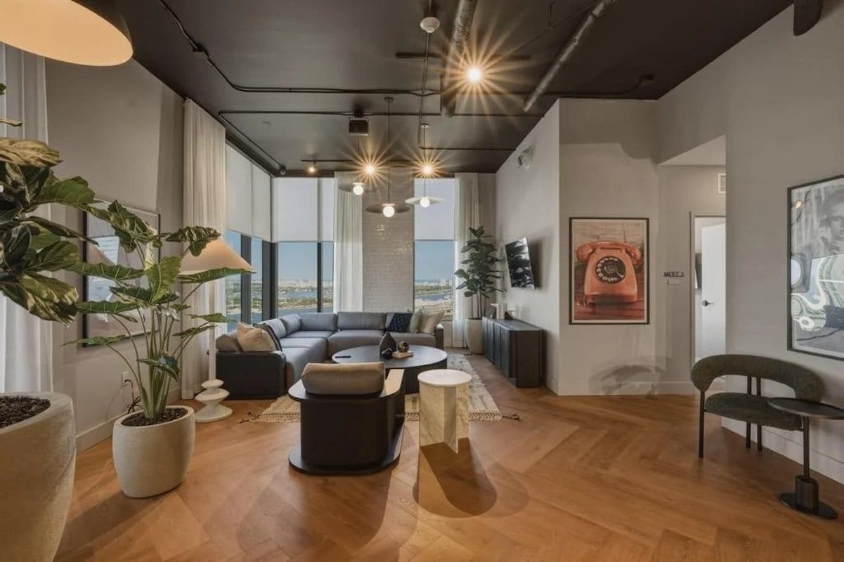 Una sala moderna con un sofá seccional, una mesa de centro redonda y un comedor con una silla. Amplias ventanas ofrecen vistas al exterior.