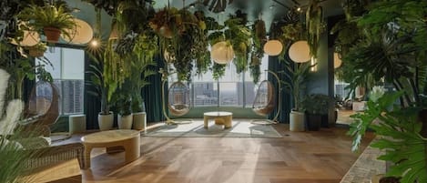 A spacious room with a high ceiling, wooden flooring, and a variety of potted plants. There are two round tables and a seating area with chairs.