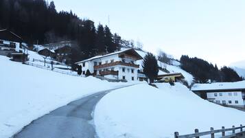 A snow-covered road leading to a multi-story building with a balcony, surrounded by trees and a mountain backdrop.
