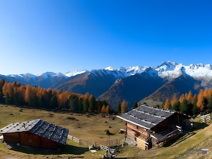 Eine Berglandschaft mit einer Holzhütte und einem kleinen Gebäude mit geneigtem Dach, umgeben von herbstlich gefärbten Bäumen und schneebedeckten Bergen.