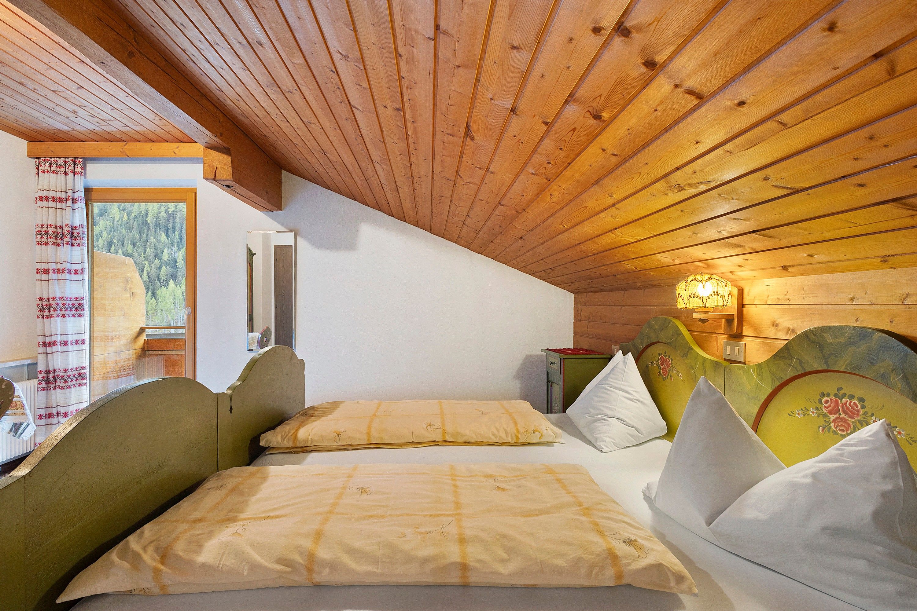 Une pièce avec un plafond en pente, un lit doté d’une tête de lit, une fenêtre donnant sur des arbres et un luminaire fixé au mur.