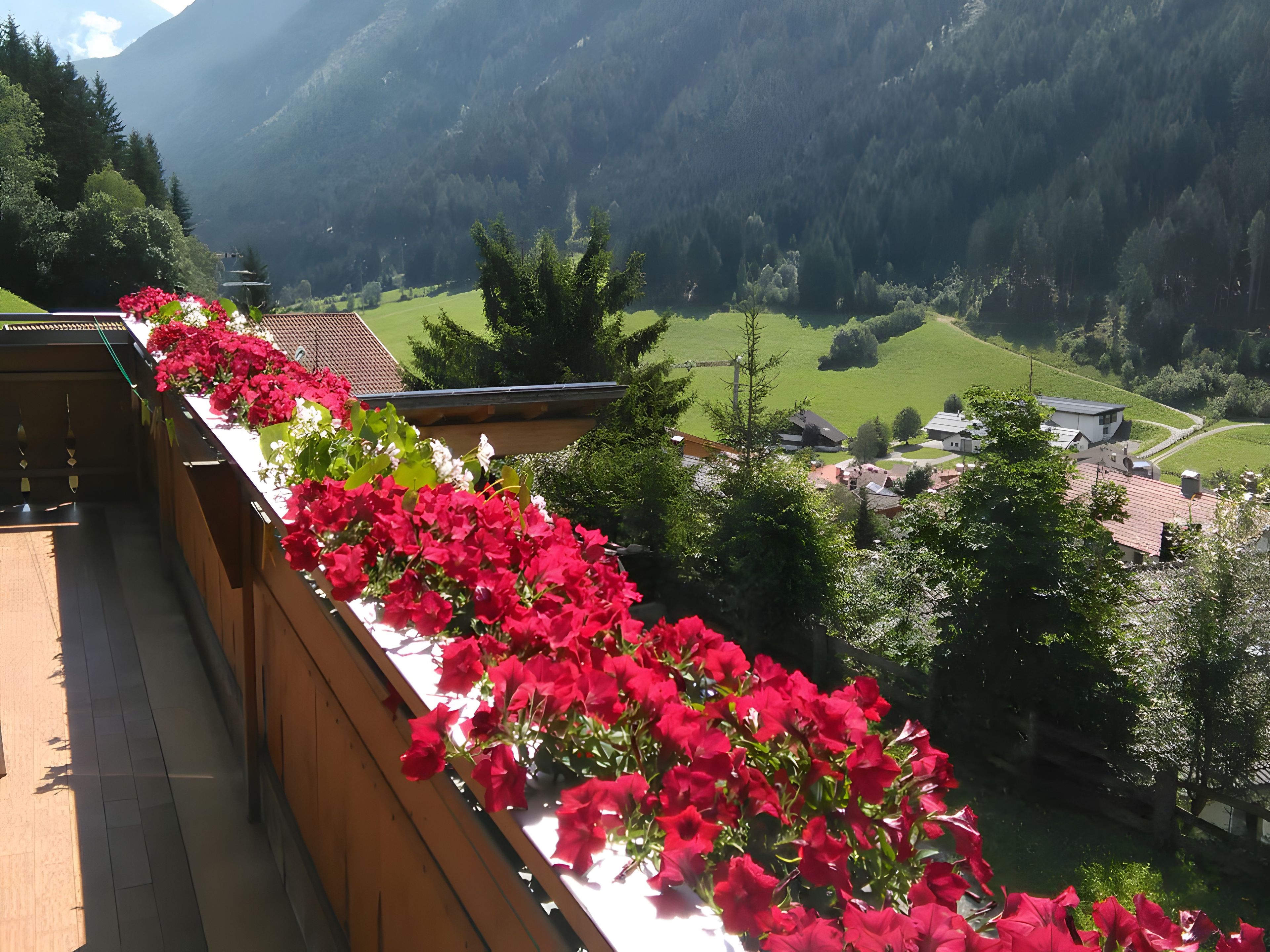 Ein Balkon mit roten Blumen, Blick auf ein Dorf und Berge im Hintergrund.