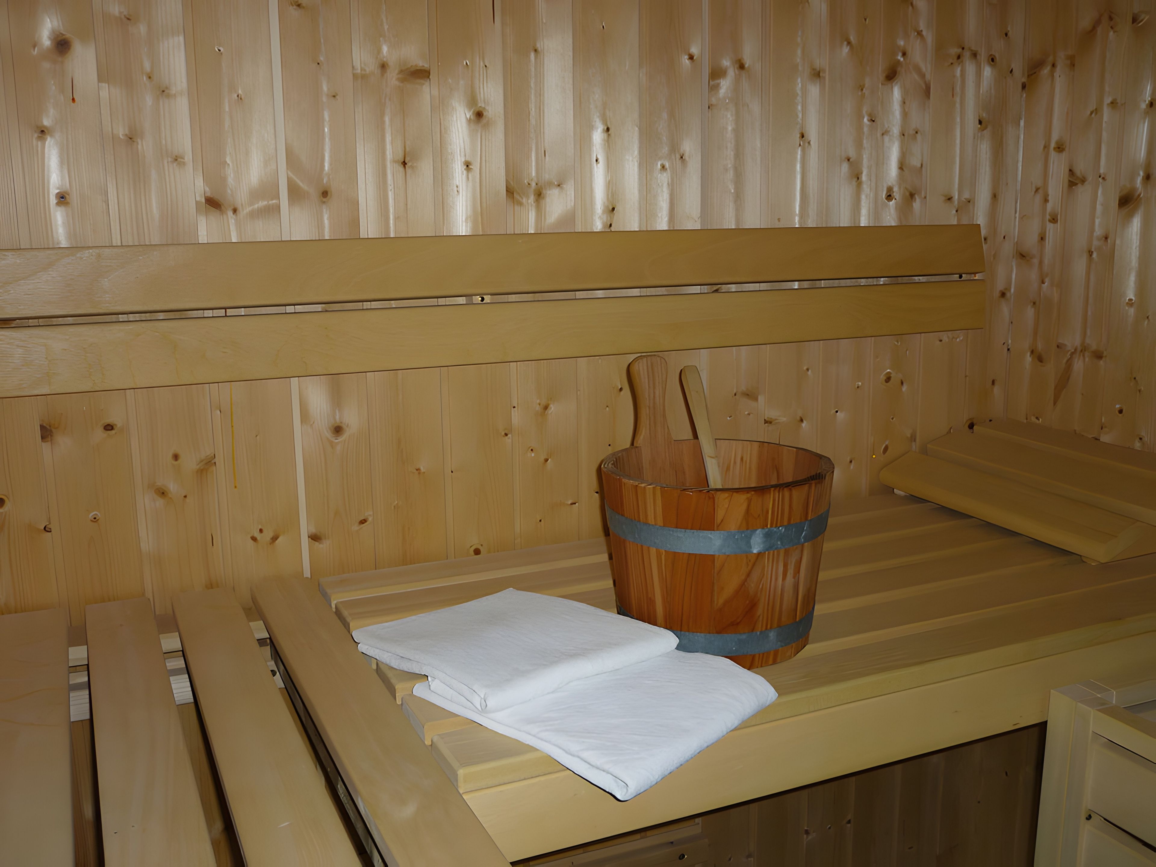 Ein Saunaraum mit Holzbänken, einem Holzwaschkübel und einem Holzklopper, sowie ein weißes Handtuch auf der Bank.