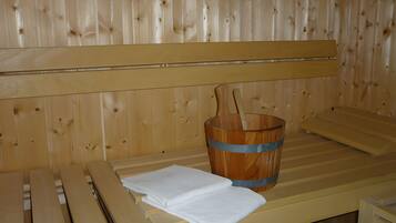 Ein Saunaraum mit Holzbänken, einem Holzwaschkübel und einem Holzklopper, sowie ein weißes Handtuch auf der Bank.